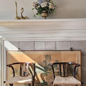 May include: Two dark wood chairs with light tan seats are in front of a large, framed Asian-style painting. Above, a white mantel holds brass swan figurines and a gold vase with flowers. A colorful, geometric wall hanging is above the mantel.