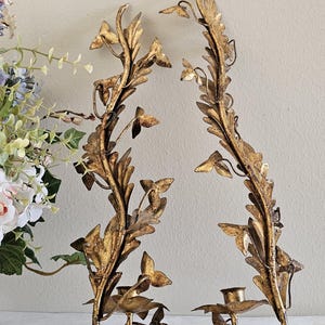 May include: Two ornate, gold-colored wall sconces with a botanical design. Each sconce features a central stem with leaf and flower details, and a small cup for holding a candle. The sconces are made of metal and have a vintage aesthetic.