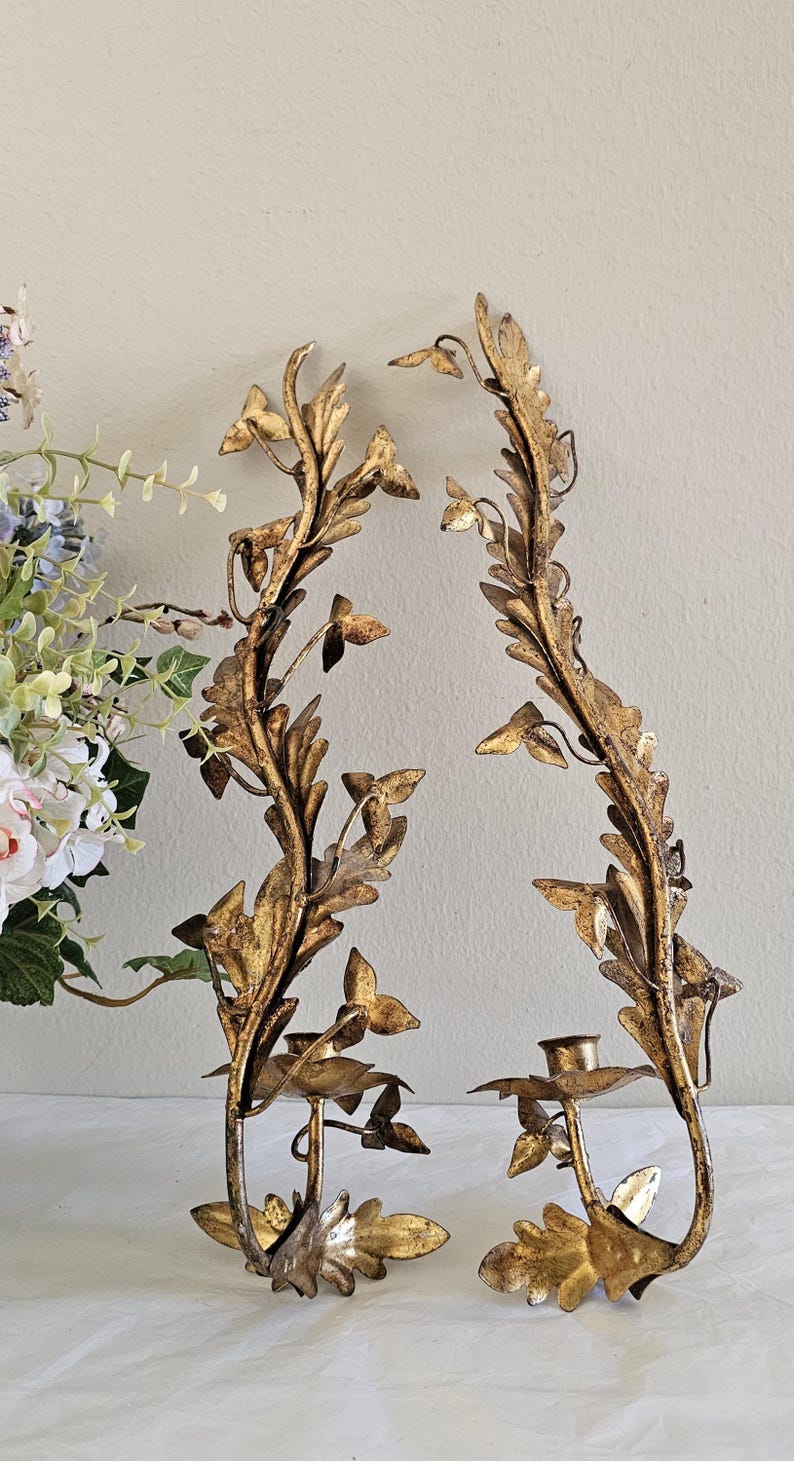 May include: Two ornate, gold-colored wall sconces with a botanical design. Each sconce features a central stem with leaf and flower details, and a small cup for holding a candle. The sconces are made of metal and have a vintage aesthetic.