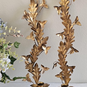 May include: Two ornate, gold-colored wall sconces with a botanical design. Each sconce features a central stem with stylized leaves and small, butterfly-shaped accents. The sconces have a candle holder at the base.