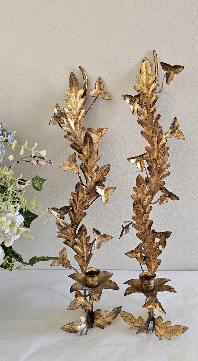 May include: Two ornate, gold-colored wall sconces with a botanical design. Each sconce features a central stem with stylized leaves and small, butterfly-shaped accents. The sconces have a candle holder at the base.