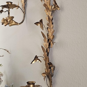 May include: Gold-colored wall sconces with a floral and leaf design. The sconces have a candle holder and are mounted on a light-colored wall. The sconces are made of metal and have a vintage aesthetic.