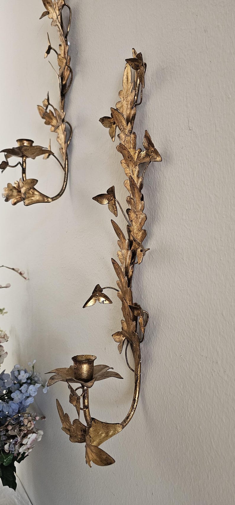 May include: Gold-colored wall sconces with a floral and leaf design. The sconces have a candle holder and are mounted on a light-colored wall. The sconces are made of metal and have a vintage aesthetic.
