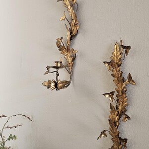 May include: Two ornate, gold-colored wall sconces with a botanical design. Each sconce features a series of leaves and small candle holders. The sconces are mounted on a light gray wall, creating a classic, elegant aesthetic.