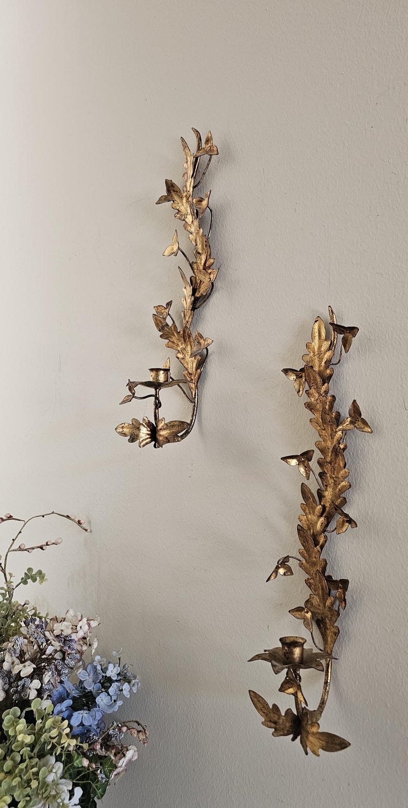 May include: Two ornate, gold-colored wall sconces with a botanical design. Each sconce features a series of leaves and small candle holders. The sconces are mounted on a light gray wall, creating a classic, elegant aesthetic.