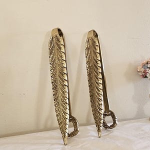 May include: Two gold-colored serving tongs with a feather design. The tongs are made of metal and have a detailed, textured pattern. The handles have a loop design. The tongs are displayed against a white background.
