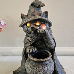 May include: A cat statue dressed as a wizard, wearing a pointed hat and cape. The cat holds a glowing orb and a cauldron. The statue has a textured, weathered finish, and the eyes glow orange. A Halloween-themed decorative item.