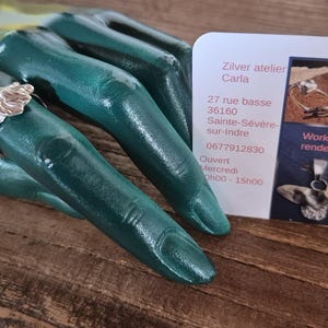 Op de afbeelding: Een zilveren ring met een bloemmotief wordt getoond op een teal handsculptuur. Een visitekaartje met de tekst "Zilver atelier Carla" en contactgegevens is ook zichtbaar.