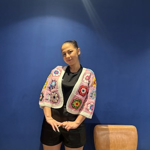 Puede incluir: Un cárdigan de ganchillo rosa, blanco y multicolor. El cárdigan presenta un estampado floral en varios colores, incluyendo rojo, amarillo y azul. La prenda se lleva sobre un top negro y se combina con unos pantalones cortos negros.