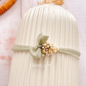 Op de afbeelding: Een lichtgroene baby haarband met een strik en kleine bloemaccenten. De haarband is gewikkeld rond een witte, geribbelde keramische vaas. De tekst "GlennWool" is op de vaas gedrukt.