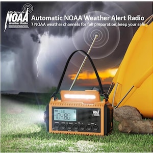May include: Orange and black NOAA weather alert radio with a digital display showing 3:30 and 104.80. The radio has a flashlight, a hand crank, and a solar panel. The radio is sitting on green grass in front of a yellow tent.