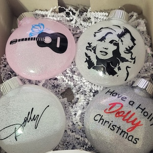 May include: Four silver glitter Christmas ornaments with different designs. One ornament features a guitar and butterfly, another has a silhouette of Dolly Parton, another has a signature, and the last one says "Have a Holly Dolly Christmas".