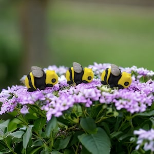 May include: Three small, yellow and black bumble bee figurines are arranged in a row on a bed of purple flowers.