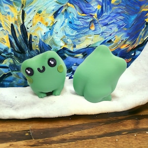 May include: Two green frog figurines. One frog has a smiling face with black eyes and blush spots. The other frog is a solid green shape. The background is a colorful painting with blue, yellow, and white brushstrokes.