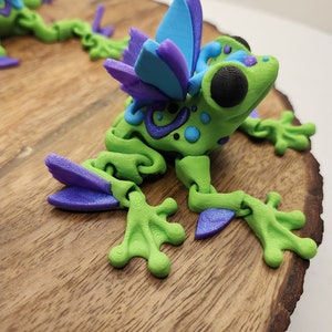 May include: A green frog figurine with purple and blue wings. The frog has black eyes and is decorated with green, blue, and purple swirls. The figurine is sitting on a wooden surface.