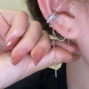 Pode incluir: Um brinco de orelha de prata com uma corrente pendurada. O brinco de orelha &eacute; decorado com pequenos strass transparentes.