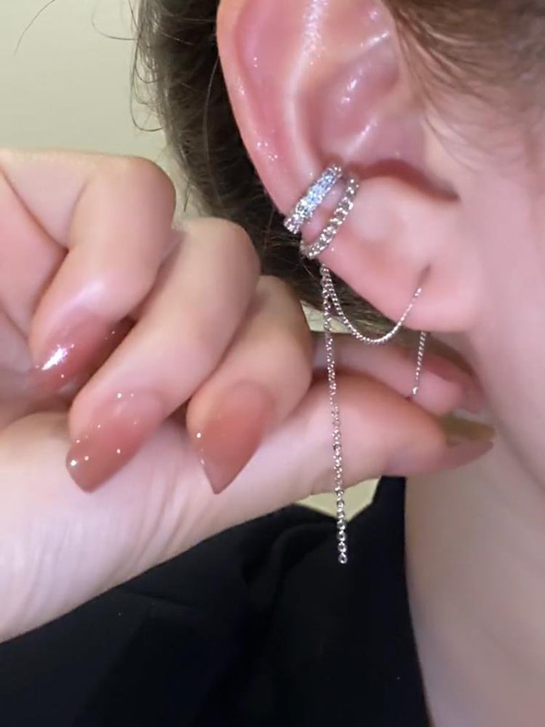 Pode incluir: Um brinco de orelha de prata com uma corrente pendurada. O brinco de orelha &eacute; decorado com pequenos strass transparentes.
