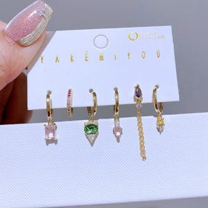 以下が含まれることがあります： さまざまな宝石のデザインが施された、6つのゴールドカラーのイヤリングのセット。 イヤリングには、ピンク、グリーン、イエローの石を使用したフープ、スタッド、ぶら下がりスタイルが含まれています。 カードには「YAKEMIYOU」と「Earring set」と書かれています。