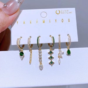 Könnte beinhalten: Ein 6-teiliges Ohrring-Set auf einer weißen Karte. Die Ohrringe sind goldfarbene Creolen mit verschiedenen baumelnden Anhängern, darunter grüne und klare Edelsteine. Auf der Karte steht "YAREMIYOU" und "6-piece Earring set".