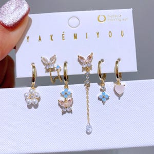 May include: A six-piece set of gold-coloured earrings. The earrings feature diverse designs, including pearls, butterflies, and heart-shaped charms. Some earrings have dangling chains and blue or clear gemstones. The card reads "YAKÉMIYOU" and "6-piece earring set."