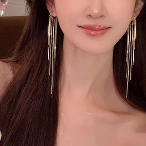 Può includere: Un paio di orecchini lunghi dorati pendenti con più catene sottili. Gli orecchini sono indossati da una persona con capelli lunghi e scuri.