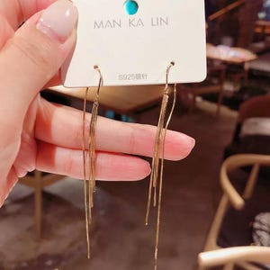Könnte beinhalten: Ein Paar goldfarbene Ohrringe mit mehreren dünnen Ketten, die von unten hängen. Die Ohrringe befinden sich auf einer weißen Karte mit dem Text "MAN KA LIN" und "S925银针".
