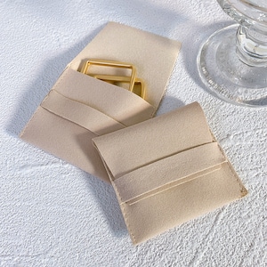 Pode incluir: Duas bolsas de feltro bege, uma com uma fivela de metal dourado no interior e a outra vazia.