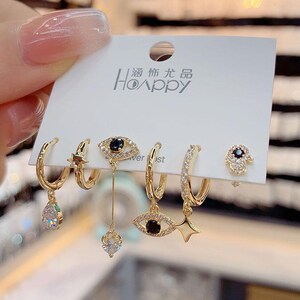 May include: A set of five gold-tone earrings with various charms, including an evil eye, a star, and a teardrop. The earrings are displayed on a white card with the text "Hoappy" in black.