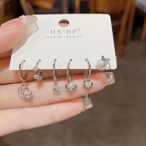 May include: A set of six silver hoop earrings with various charms, including a crescent moon, a teardrop, a star, and a heart. The charms are adorned with clear and blue crystals. The earrings are displayed on a white card with the text "HS.DP® FASHION JEWELRY".