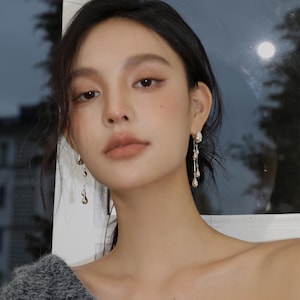 Może przedstawiać: Kobieta z długimi czarnymi włosami w srebrnych kolczykach z wiszącymi ozdobami w kształcie łez. Kolczyki są długie i delikatne, a ozdoby są małe i błyszczące.