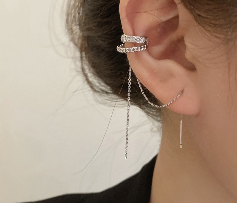 Pode incluir: Um brinco de argola prateado com uma corrente pendurada. A argola &eacute; decorada com pequenos strass transparentes. A corrente est&aacute; presa a um pequeno brinco de press&atilde;o prateado.
