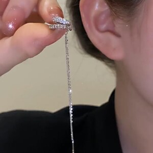 Pode incluir: Um brinco de argola prateado com um pequeno design de argola dupla cravejado com strass transparentes. O brinco tem uma corrente longa e fina que pende da argola.