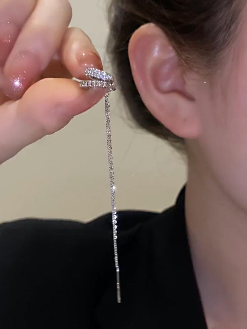 Pode incluir: Um brinco de argola prateado com um pequeno design de argola dupla cravejado com strass transparentes. O brinco tem uma corrente longa e fina que pende da argola.