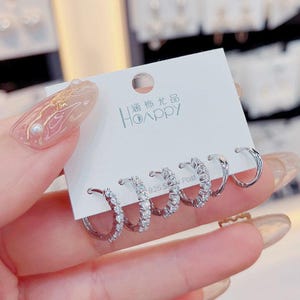 Puede incluir: Cinco pares de pendientes de aro de plata con piedras de circonita cúbica transparentes. Los pendientes se muestran en una tarjeta blanca con el texto "Happy" en chino e inglés.