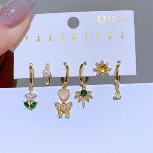 May include: A six-piece earring set featuring gold-toned hoops and studs with various charms. Charms include a flower, a pearl, a heart and butterfly, a sunburst, and a teardrop. The card reads "YAKEN MIYOU" and "6-piece Earring set".