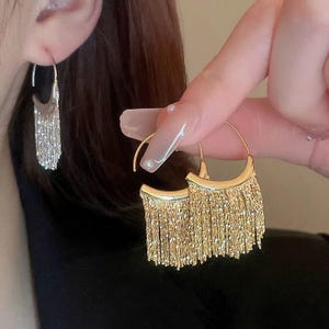 May include: A pair of gold hoop earrings with dangling chains. The earrings are made of a thin, delicate chain that hangs down from the hoop. The chains are about 5 cm long and have a fringe at the bottom.