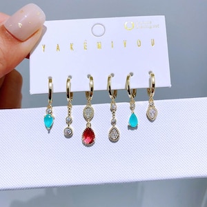 May include: A collection of six gold-tone hoop earrings, each with a different dangling charm. The charms feature turquoise and clear teardrop stones, and a red oval gem. The earrings are presented on a white card with the text "YAKEMIYOU".