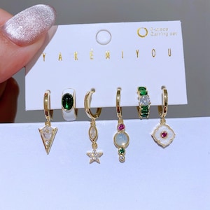 May include: A 6-piece earring set on a white card. The earrings are gold-toned hoops with various charms, including a white and green stone, a star, and a geometric design. The text "YAKEMIYOU" and "6-Piece Earring set" are visible.