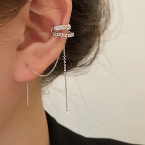 Może przedstawiać: Srebrny kolczyk typu cuff z wiszącą łańcuszkiem. Kolczyk ozdobiony jest małymi, przezroczystymi cyrkoniami. Łańcuszek jest przymocowany do małego srebrnego kolczyka.