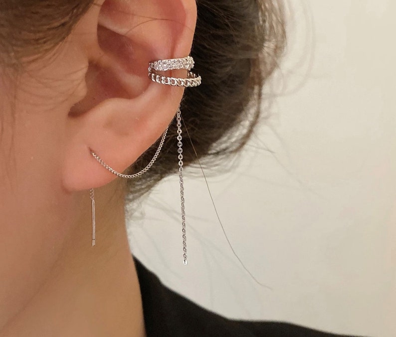Pode incluir: Um brinco de argola prateado com uma corrente pendurada. A argola &eacute; adornada com pequenos strass transparentes. A corrente est&aacute; presa a um pequeno brinco de press&atilde;o prateado.
