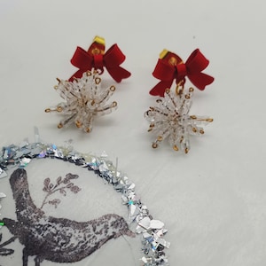 May include: A pair of earrings with a clear beaded snowflake design and red bows. The earrings are made with gold-toned metal.