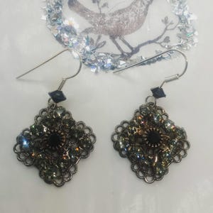 May include: A pair of ornate, filigree earrings. Each earring features a dark central stone surrounded by smaller, iridescent gems. The earrings have a dark metal finish and are suspended from silver-colored ear wires.