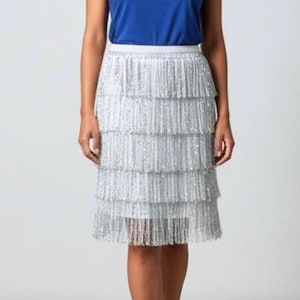 May include: A woman wearing a blue t-shirt with the text "THE FRINGE" in white, paired with a silver fringe skirt. The skirt has multiple layers of fringe. The outfit is suitable for a party or special event.