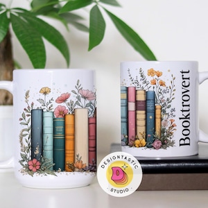 May include: Two white ceramic mugs with floral and book designs. One mug has the word "Booktrovert" printed vertically. The mugs are decorated with colorful books and flowers, perfect for book lovers.