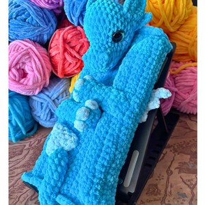 Peut inclure: Un jouet dragon en crochet bleu clair, avec un œil noir et des détails blancs. Le dragon est posé sur un support noir, avec des pelotes de laine colorées en arrière-plan. Les couleurs de la laine sont rose, bleu, rouge et jaune.