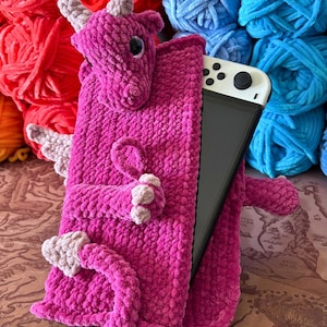 Op de afbeelding: Een felroze gehaakte draak-thema hoes voor een handheld game-apparaat. De hoes heeft een drakenkop, hoorns, vleugels en ledematen. Het apparaat is gedeeltelijk zichtbaar. Gekleurd garen in rood, blauw en oranje is op de achtergrond.