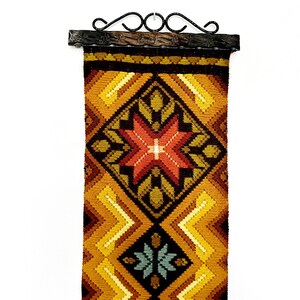 Könnte beinhalten: Ein gewebter Wandbehang mit einem geometrischen Muster in Braun-, Gelb-, Orange-, Rot- und Grüntönen. Der Behang hat ein zentrales Sternendesign und ist mit einer schwarzen Metallstange gerahmt.