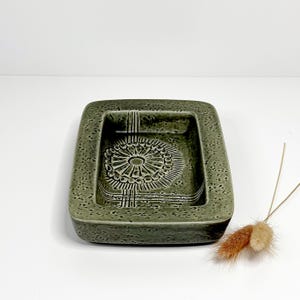 May include: A square, olive green ceramic ashtray with a textured surface. The ashtray features a central, circular design with radiating lines and a raised border. The design is reminiscent of a stylized flower or sun. The ashtray is a vintage piece.