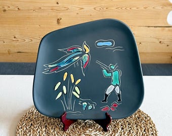 May include: A dark teal ceramic plate with a unique shape, featuring a hand-painted hunting scene. The design includes a hunter, bird, and foliage in shades of teal, blue, yellow, and red. The plate is displayed on a small, dark red stand.