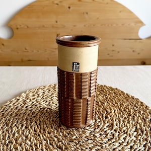 May include: A brown ceramic vase with a beige top and a black label that says "J.ie Verk. Stad." The vase is sitting on a woven brown mat.
